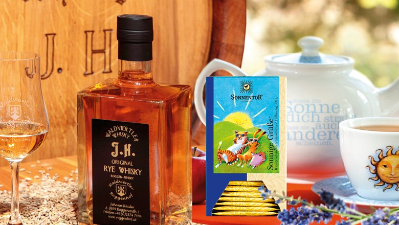 Der Weg der Kräuter und Gewürze, die Faszination Whisky, © Sonnentor und Whiskydestillerie Eine Flasche Waldviertler Rye Whisky neben einem Glas, Teepackung und Teekanne auf einem Tisch mit Lavendel und Getreide.
