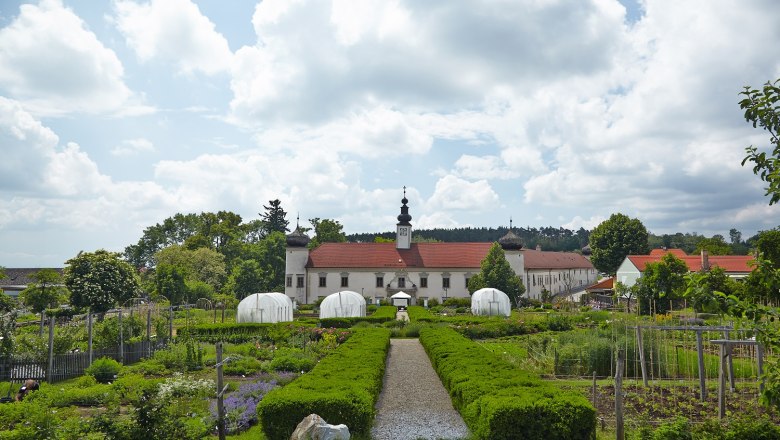 Arche Noah, © Rupert Pessl Ein gepflegter Garten mit Gewächshäusern vor einem historischen Gebäude mit rotem Dach und Turm, umgeben von Bäumen und Wolken am Himmel.