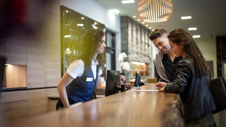 Sole Felsen Welt, © Sole Felsen Welt Ein Paar checkt an der Rezeption eines Hotels ein, während eine Rezeptionistin ihnen hilft.