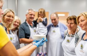 Eine Gruppe von Menschen betrachtet einen frisch hergestellten Käse, der von einer Person mit blauen Handschuhen gehalten wird.