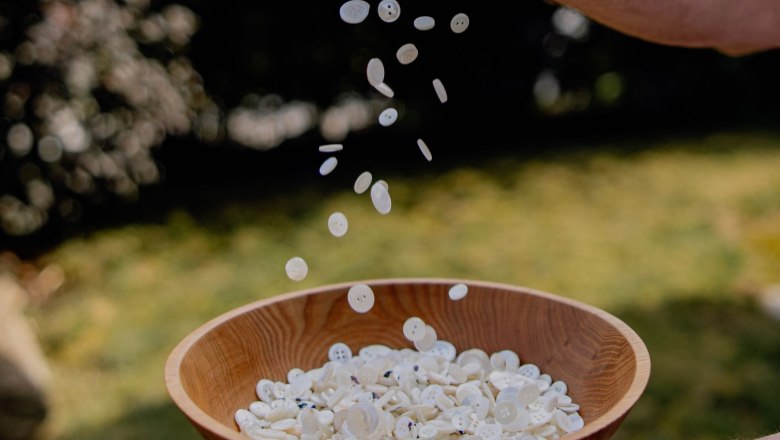Eine Hand h&auml;lt eine Holzschale mit wei&szlig;en Kn&ouml;pfen, w&auml;hrend weitere Kn&ouml;pfe von oben hineinfallen.