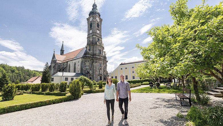 Ein Paar spaziert im Garten des Stifts Zwettl, im Hintergrund die Kirche und ein blauer Himmel.
