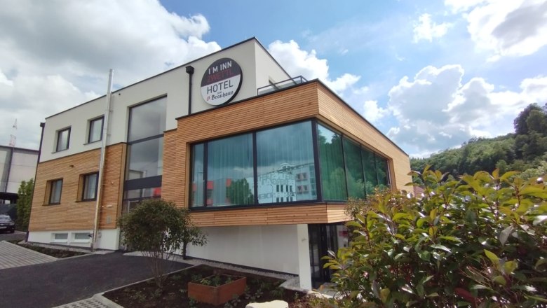 Modernes Hotelgeb&auml;ude mit Holzverkleidung und gro&szlig;en Fenstern, umgeben von Pflanzen, unter blauem Himmel mit Wolken.
