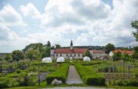 Ein gepflegter Garten mit Gew&auml;chsh&auml;usern vor einem historischen Geb&auml;ude mit rotem Dach und Turm, umgeben von B&auml;umen und Wolken am Himmel.