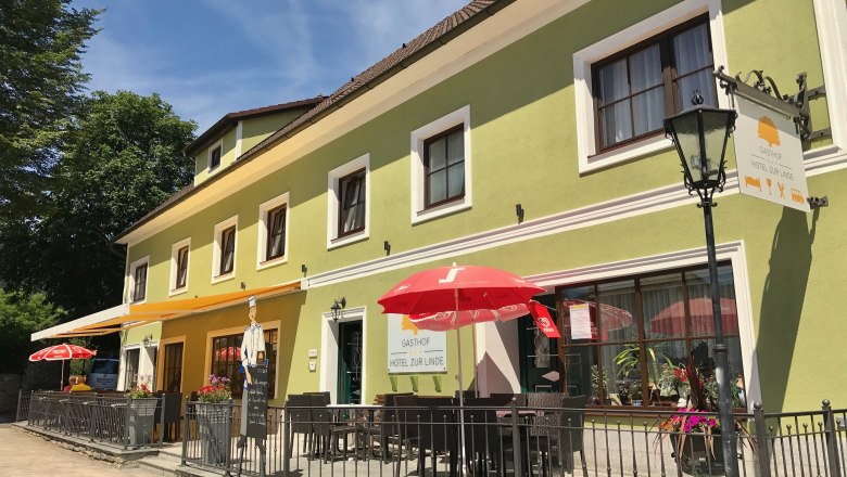 Ein gr&uuml;ner Gasthof mit Terrasse und roten Sonnenschirmen, beschriftet mit 'Gasthof zur Linde'.