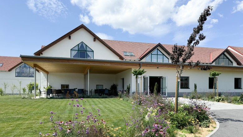 Ein modernes Gebäude mit rotem Dach, umgeben von einem gepflegten Garten mit bunten Blumen und Rasen, unter blauem Himmel.