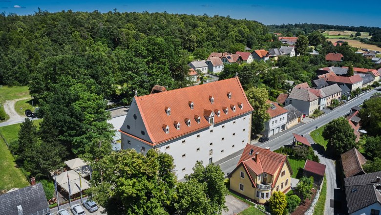Luftaufnahme eines großen Gebäudes mit rotem Dach, umgeben von Bäumen und kleineren Häusern in einer ländlichen Gegend.