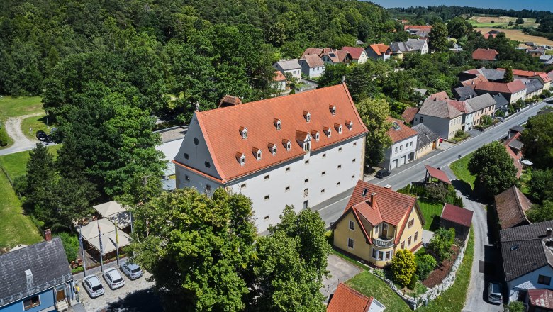 Luftaufnahme eines großen Gebäudes mit rotem Dach, umgeben von Bäumen und kleineren Häusern in einer ländlichen Gegend.